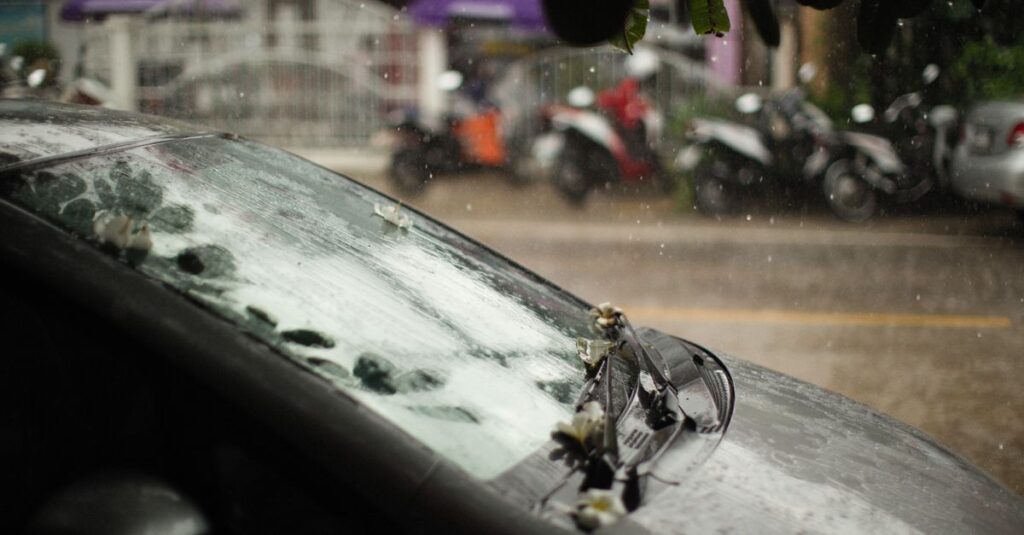 Close-up view of a black car windshield with raindrops on a rainy day streetscape.